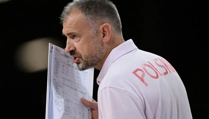Trener Nikola Grbić wpatrzony w dokumenty, z napisem "POLSKA" na koszulce. Może analizuje statystyki, myśląc o swojej żonie.