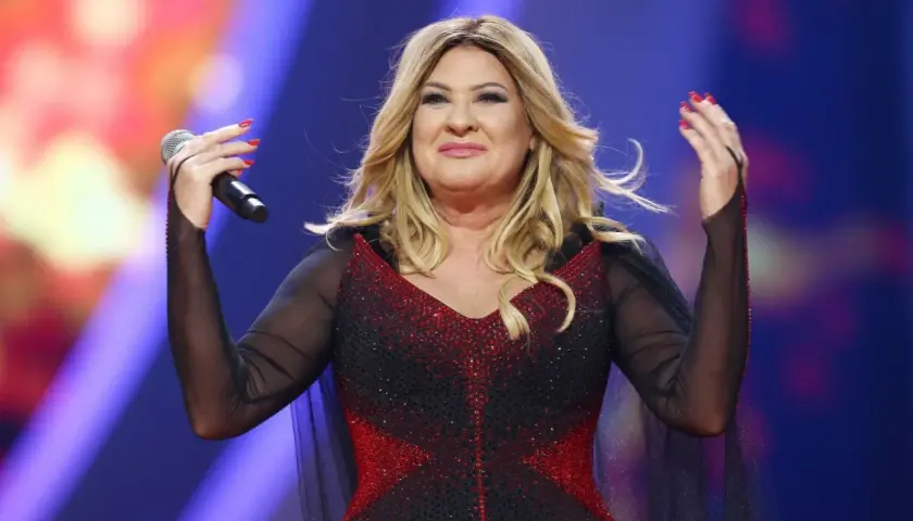 Beata Kozidrak na scenie, w błyszczącej sukni, z mikrofonem w dłoni. Jej wzrost dodaje jej scenicznej prezencji.