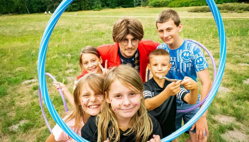 Rafał Brzoska z córkami i przyjaciółmi, wesołe dzieci z brokatem na twarzach, bawią się na trawie z hula-hop.