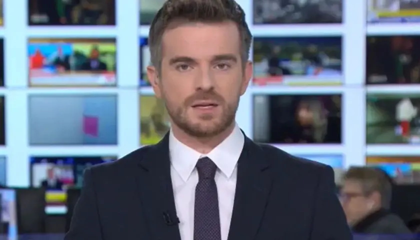 Łukasz Jedliński, dziennikarz, w garniturze i krawacie, na tle ekranów informacyjnych. Jego życiorys to historia kariery w mediach.