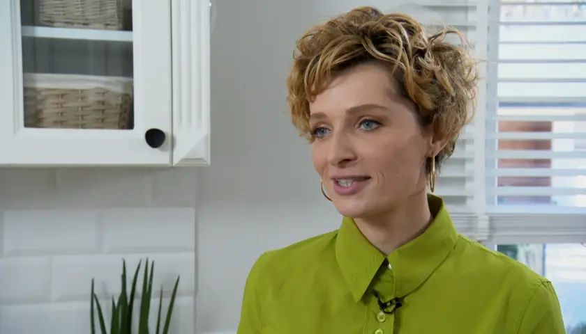 Katarzyna Jaroszyńska, wiek nieznany, w zielonej koszuli, z kręconymi blond włosami, uśmiecha się. W tle biała kuchnia.