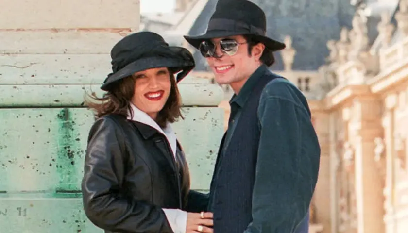 Lisa Marie Presley i jej mąż Michael Jackson w kapeluszach i okularach przeciwsłonecznych, uśmiechnięci na tle budowli.