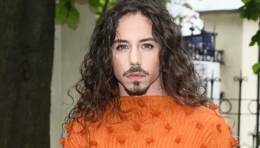 Michał Szpak w pomarańczowej bluzce z falbanami, z burzą loków i charakterystyczną bródką. Jego wiek nie wpływa na jego styl.