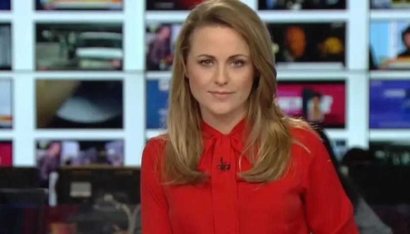 Marta Kłos, w czerwonej bluzce, na tle ekranów informacyjnych. Jej biografia to historia kariery w mediach.
