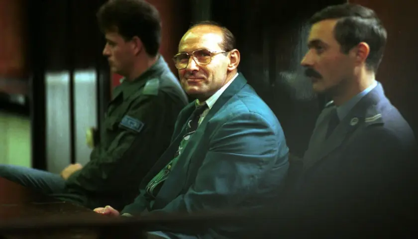 Trzech mężczyzn w garniturach siedzi w sali sądowej. Jeden z nich, w okularach, wygląda jak pershing gangster.