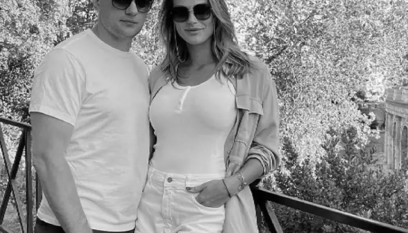 Aryna Sabalenka z partnerem, uśmiechnięci, w okularach przeciwsłonecznych, na tle drzew i architektury.