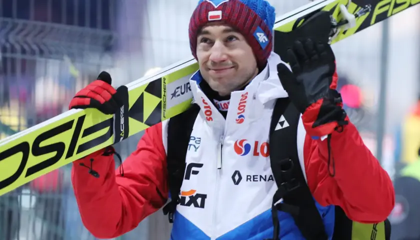 Uśmiechnięty Andrzej Stoch Jr. z nartami na ramieniu, gotowy do skoku.