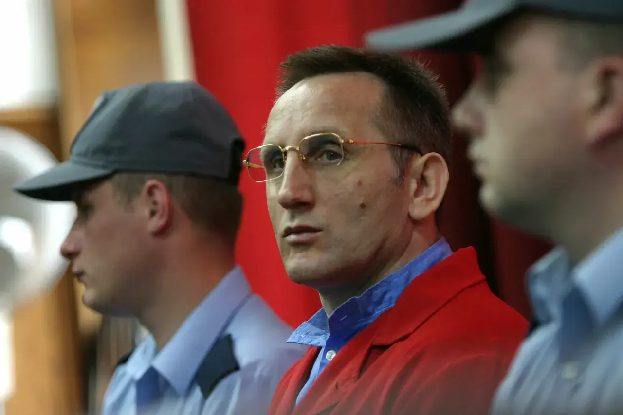 Słowik gangster w czerwonej marynarce, otoczony przez ochroniarzy w niebieskich koszulach i czapkach.