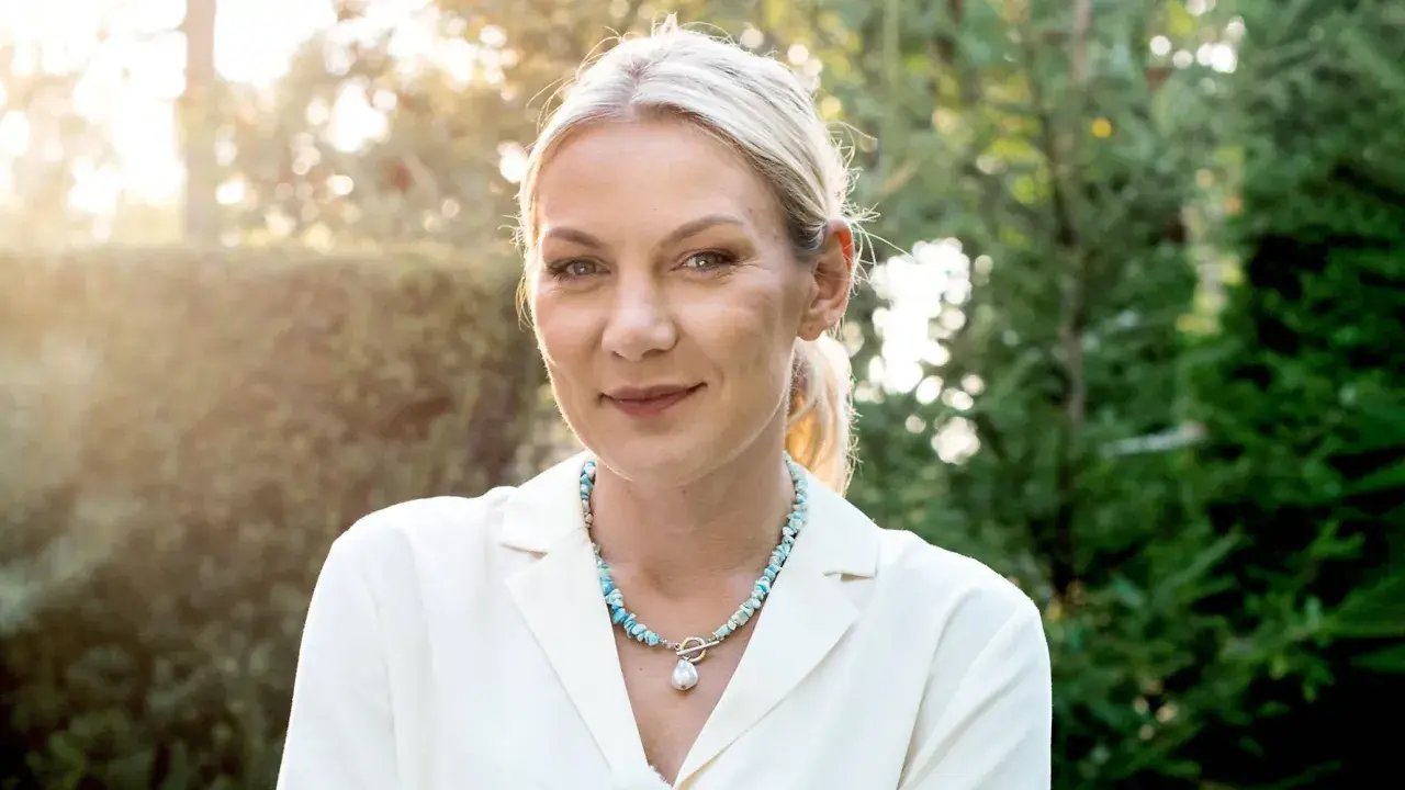 Uśmiechnięta Martyna Kupczyk w białej marynarce i naszyjniku z pereł. W tle zielony ogród.