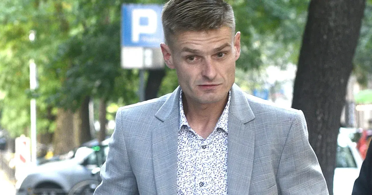 Tomasz Komenda w szarym garniturze i wzorzystej koszuli, z widocznym znakiem P. Jego wiek jest trudny do określenia na pierwszy rzut oka.