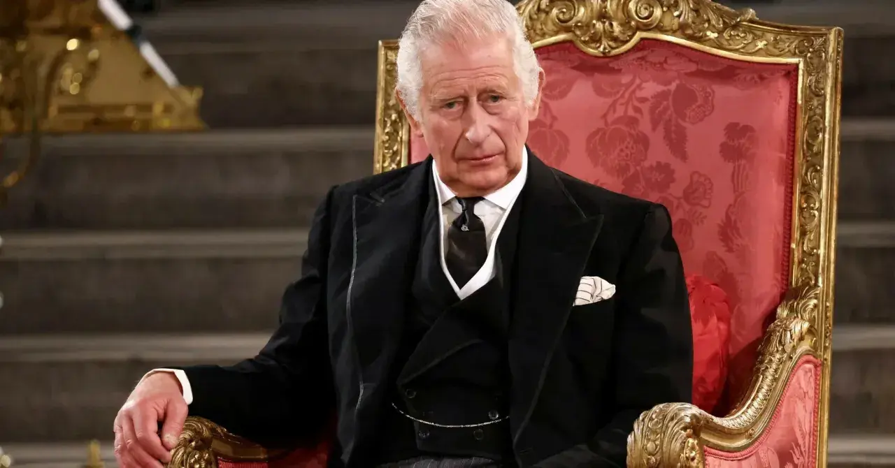 Król Karol w wieku siedemdziesięciu lat, w eleganckim czarnym garniturze, siedzi na ozdobnym tronie.