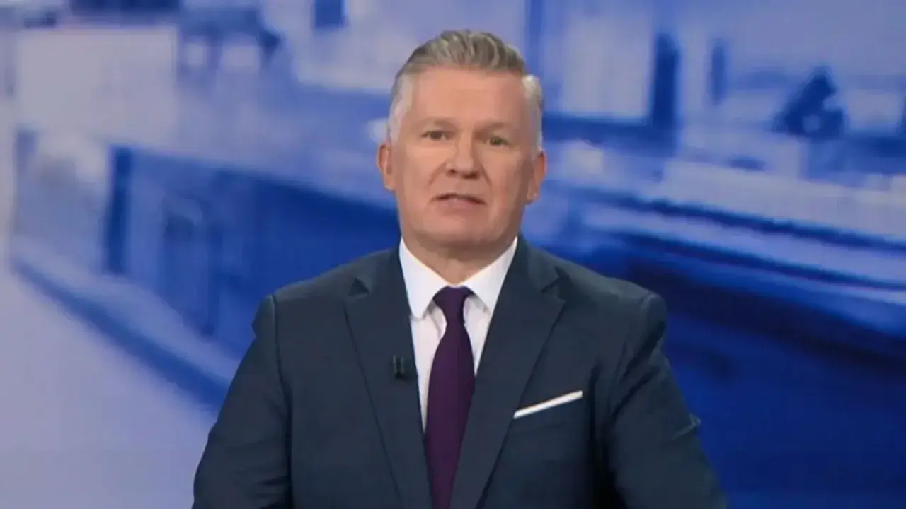 Jeden z prezenterów Fakty TVN w granatowym garniturze i bordowym krawacie, na tle niebieskiej grafiki.