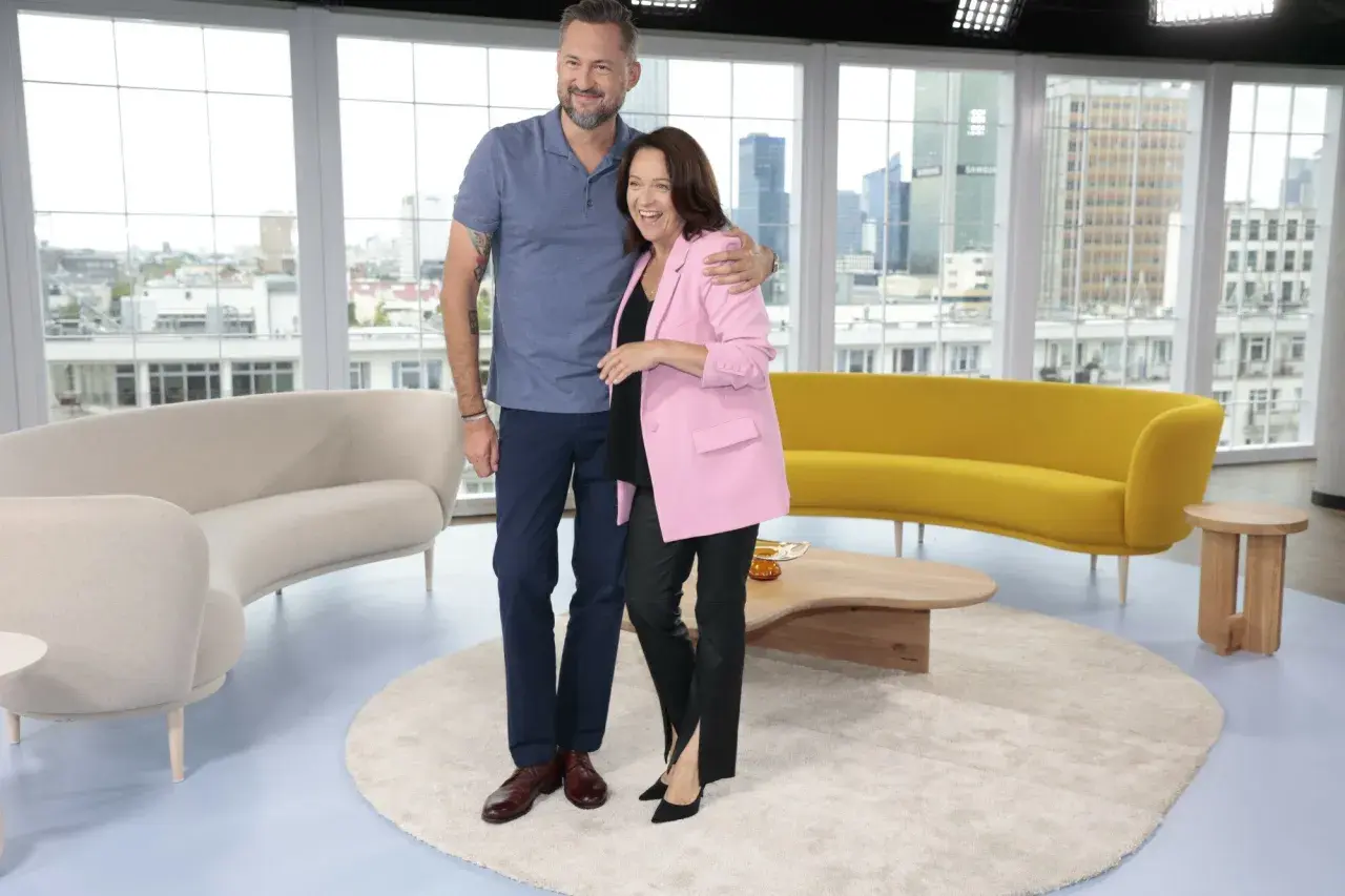 Ewa Drzyzga z mężem w studiu telewizyjnym, uśmiechnięci, na tle okien z widokiem na miasto.