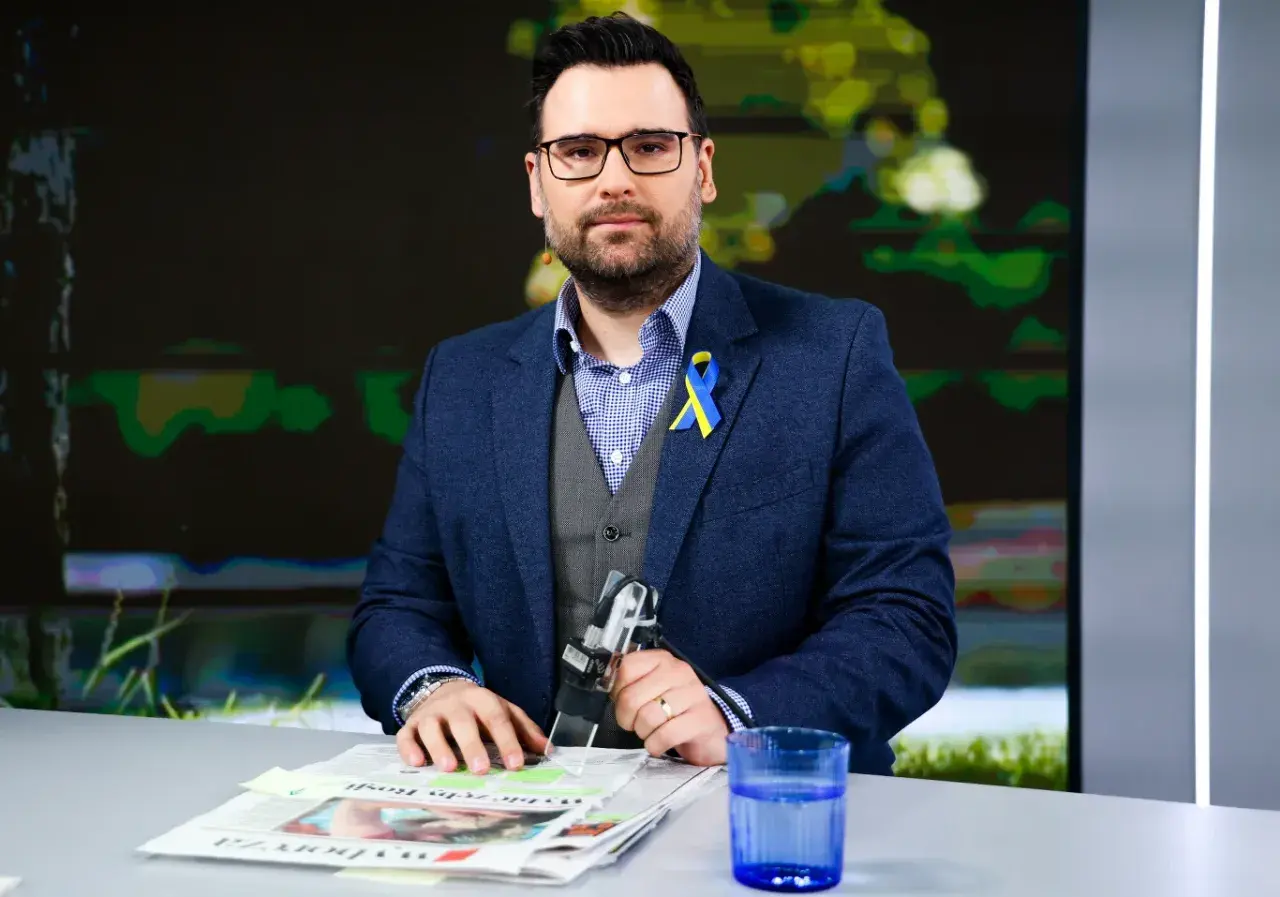 Michał Szajder, wiek nieznany, w garniturze z przypinką w barwach Ukrainy, siedzi przy stole z gazetami i szklanką wody.