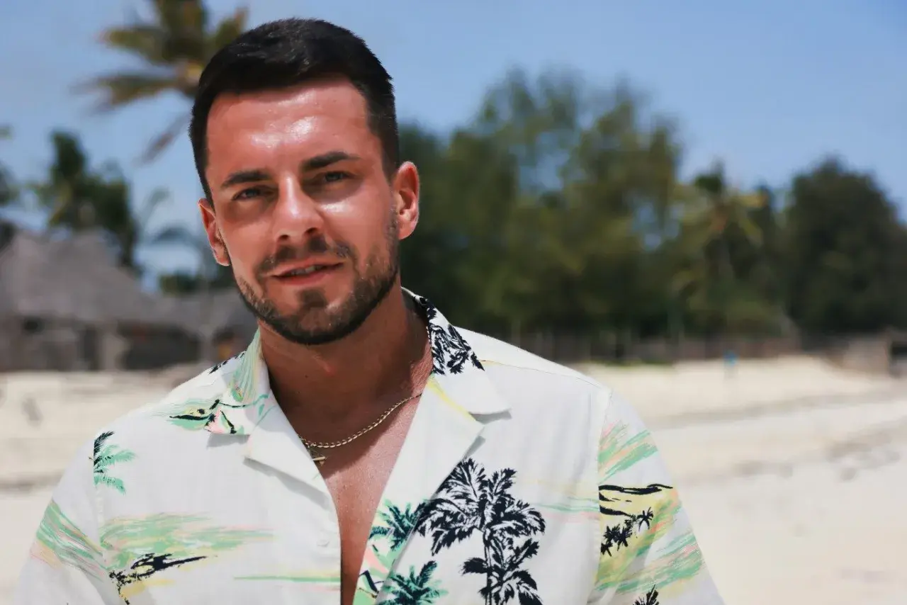 Basia z Hotelu Paradise na plaży, w hawajskiej koszuli, z palmami w tle.