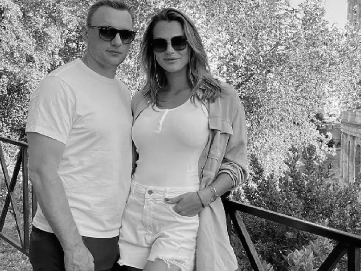Aryna Sabalenka z partnerem, uśmiechnięci, w okularach przeciwsłonecznych, na tle drzew i architektury.