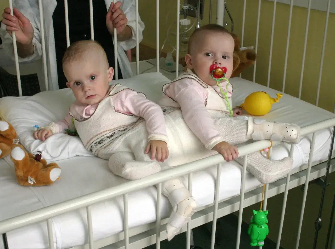 Dwie urocze bliźniaczki syjamskie z Janikowa w łóżeczku szpitalnym, otoczone pluszakami i zabawkami.