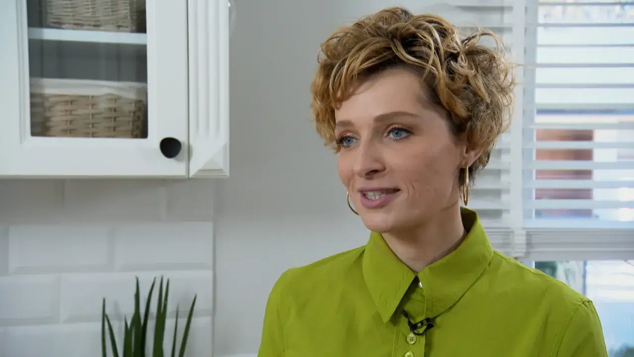 Katarzyna Jaroszyńska, w wieku około 30 lat, w zielonej koszuli, uśmiecha się. W tle biała kuchnia z wiklinowymi koszami.