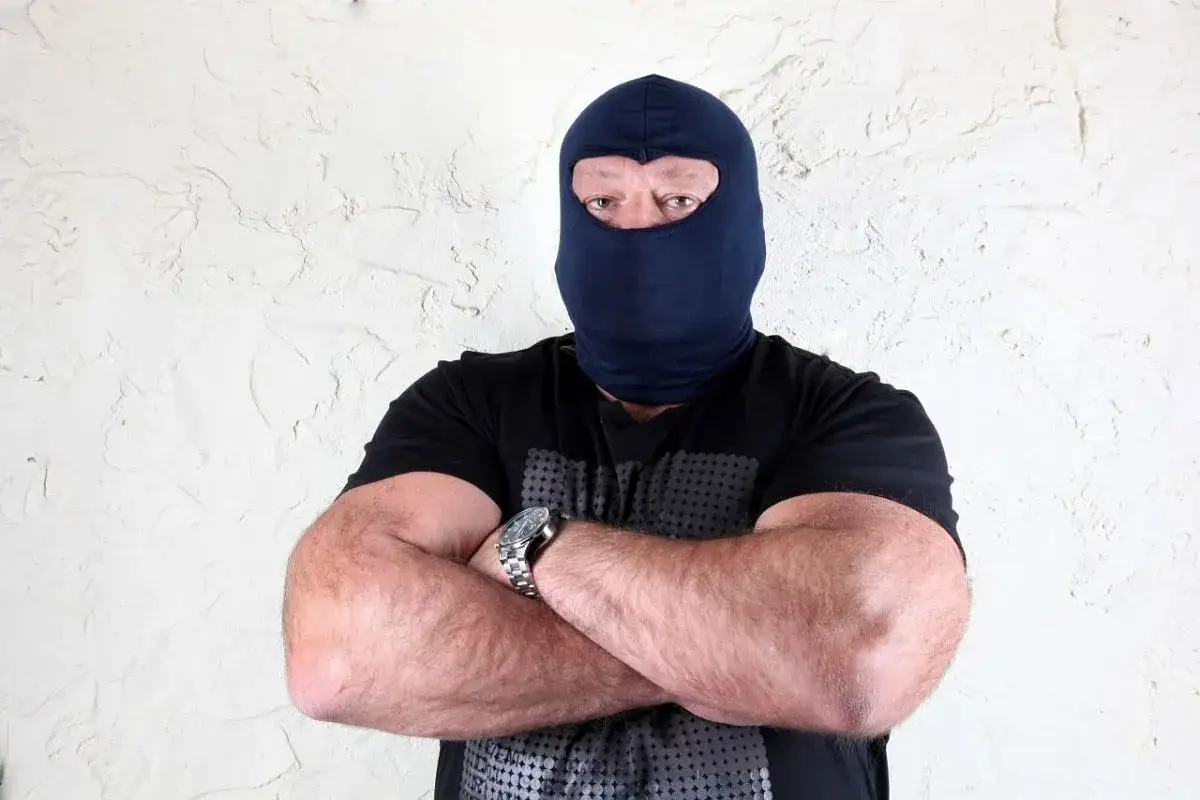 Masywny mężczyzna w kominiarce, z rękami skrzyżowanymi na piersi, wygląda jak groźny gangster.