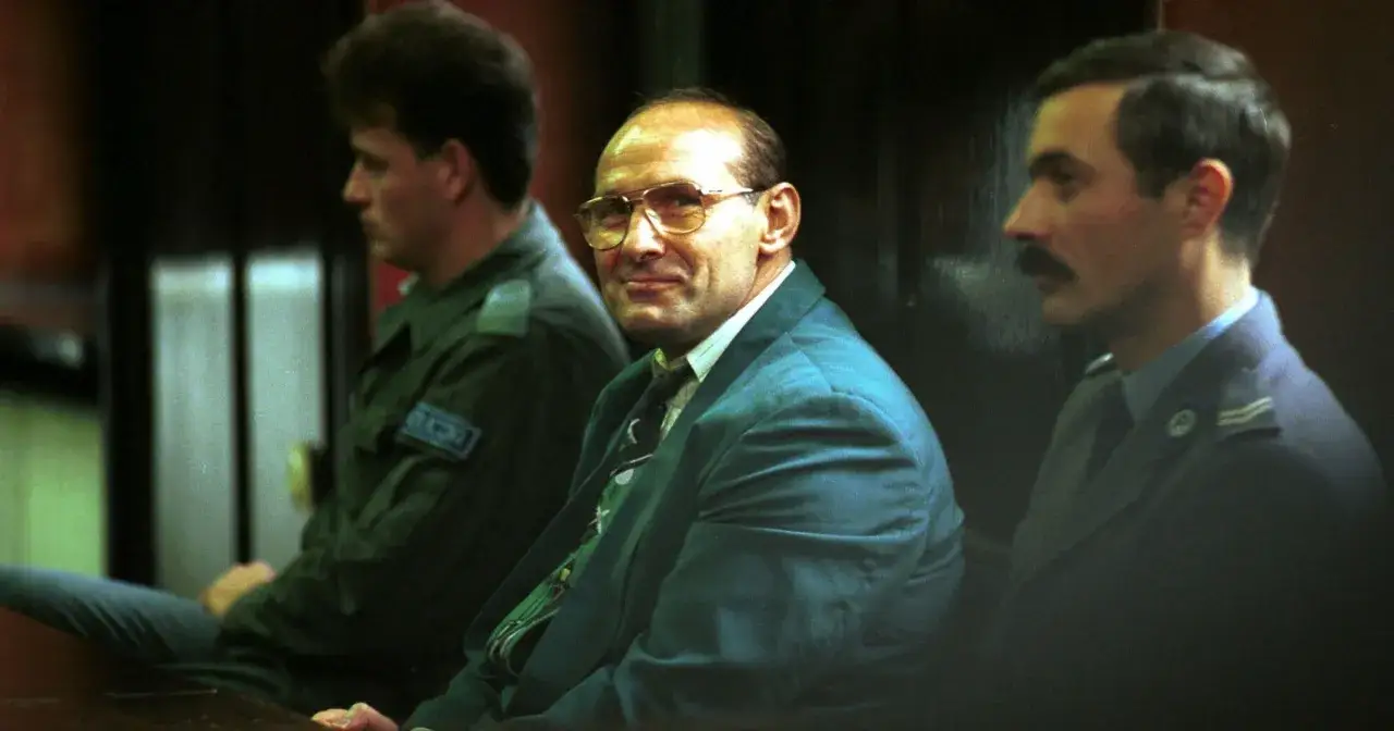 Uśmiechnięty mężczyzna w okularach, ubrany w garnitur, siedzi obok innych mężczyzn. Wygląda jak pershing gangster.
