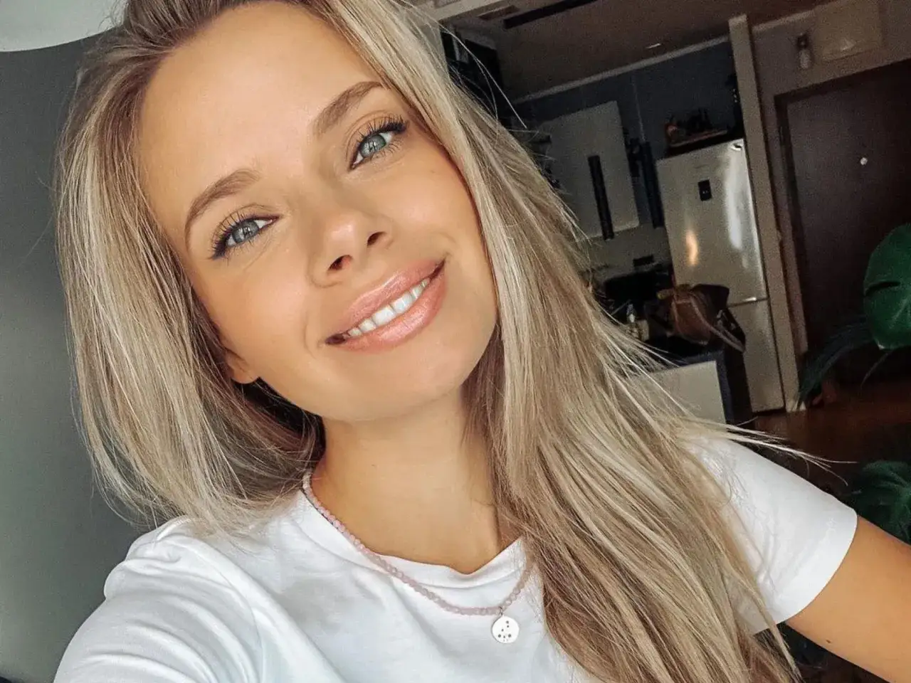 Uśmiechnięta blondynka z niebieskimi oczami, w białej koszulce, z naszyjnikiem.
