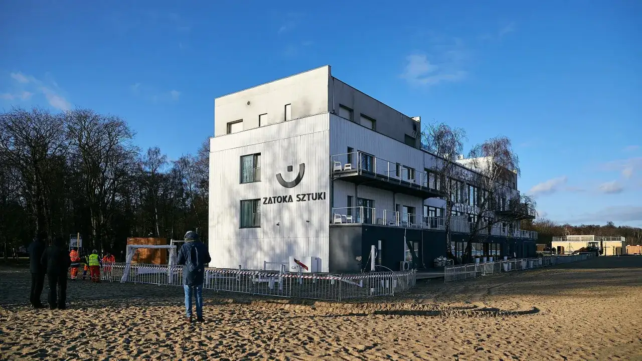 Budynek Zatoki Sztuki, gdzie toczyła się afera, na piaszczystym terenie. W tle drzewa i niebo.