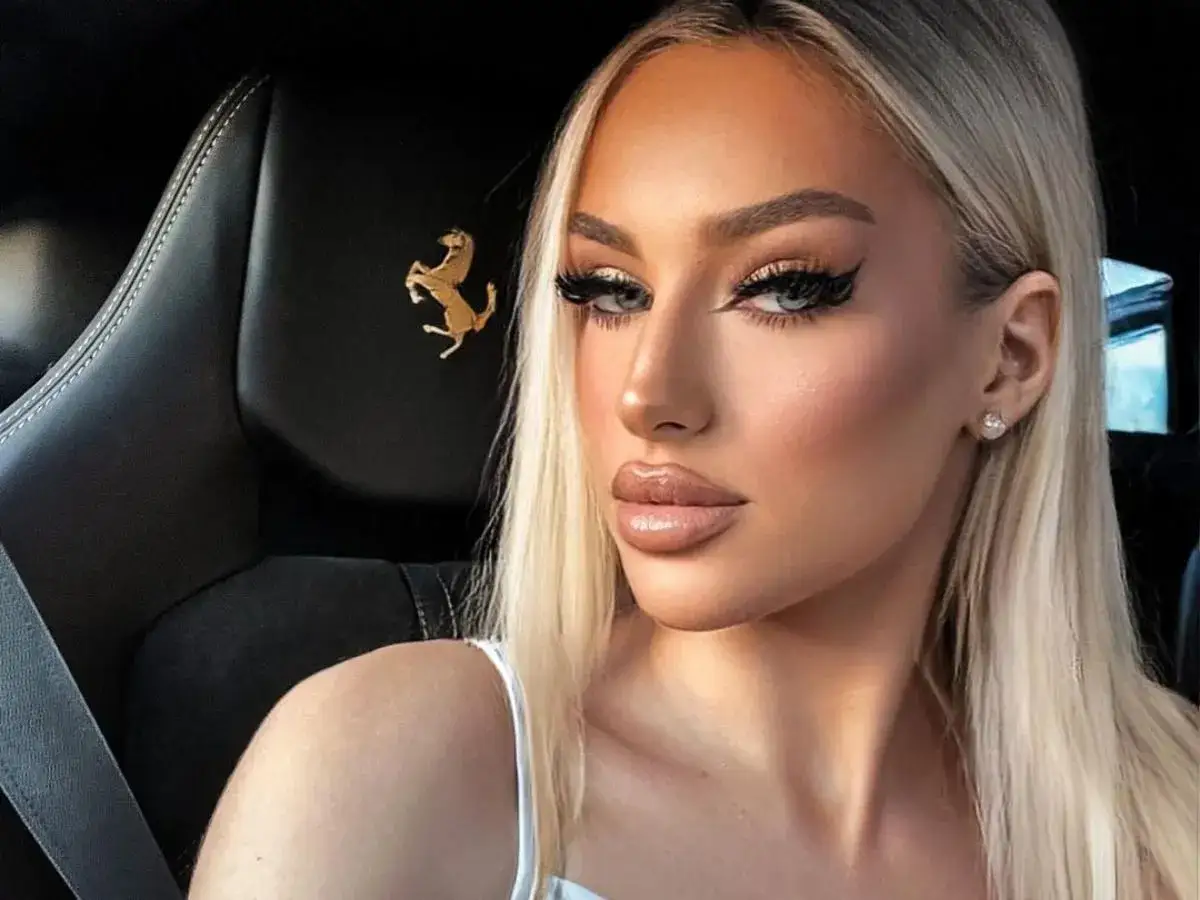 Caroline Derpienski w sportowym aucie, z charakterystycznym logo Ferrari. Jej wiek nie jest widoczny, ale makijaż sugeruje młodość.