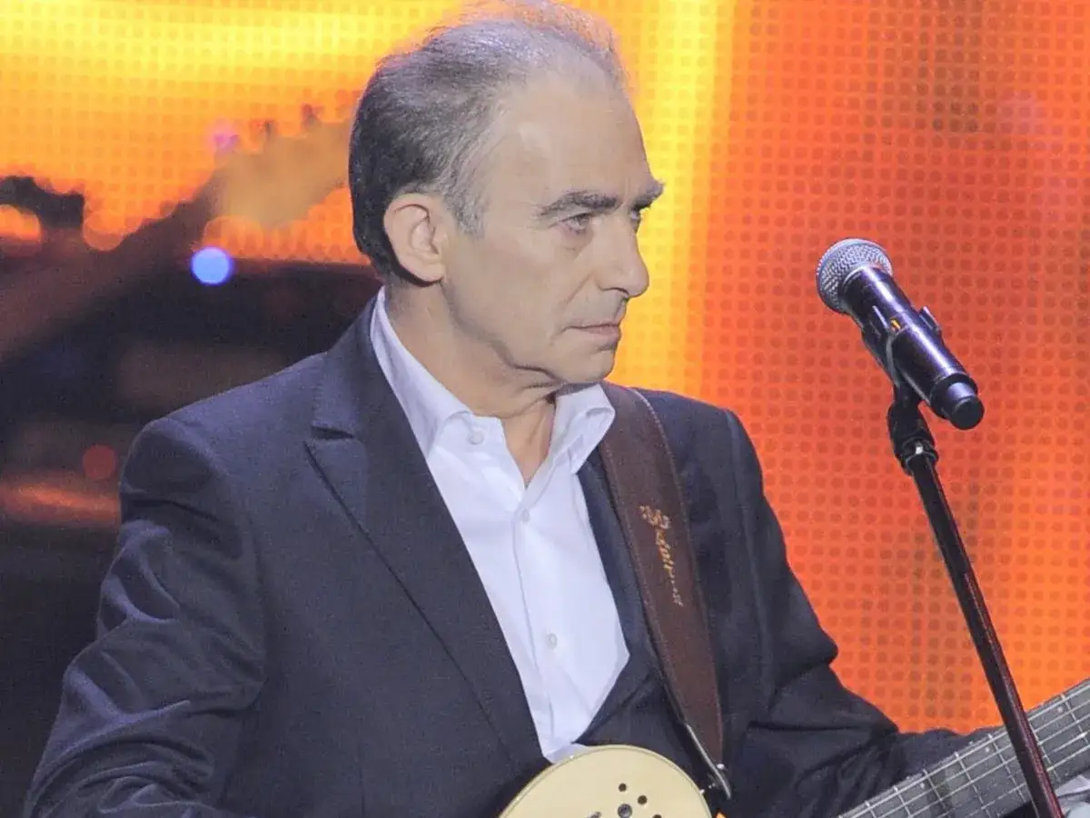 Wspomnienie Seweryna Krajewskiego, który nie żyje. Artysta w garniturze i z gitarą na scenie, przed mikrofonem.