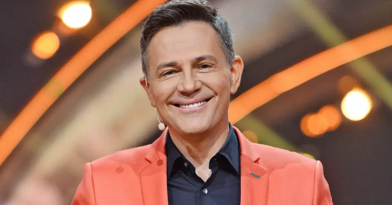 Krzysztof Ibisz w pomarańczowej marynarce, uśmiechnięty. W tle pomarańczowe światła.