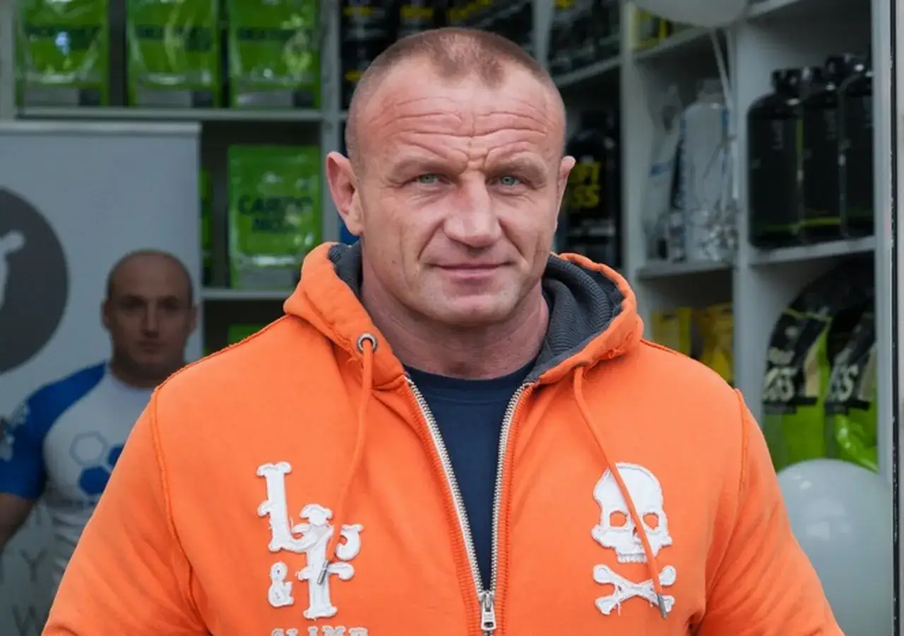 Mariusz Pudzianowski, znany z siły i determinacji, w pomarańczowej bluzie. Jego wiek nie przeszkadza mu w osiąganiu kolejnych sukcesów.