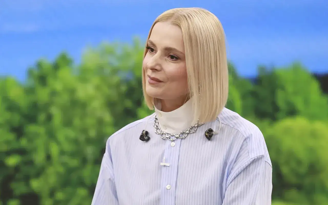 Halina Mlynkova, wiek nieznany, w błękitnej koszuli i srebrnym naszyjniku, z blond włosami, na tle zieleni i błękitu.