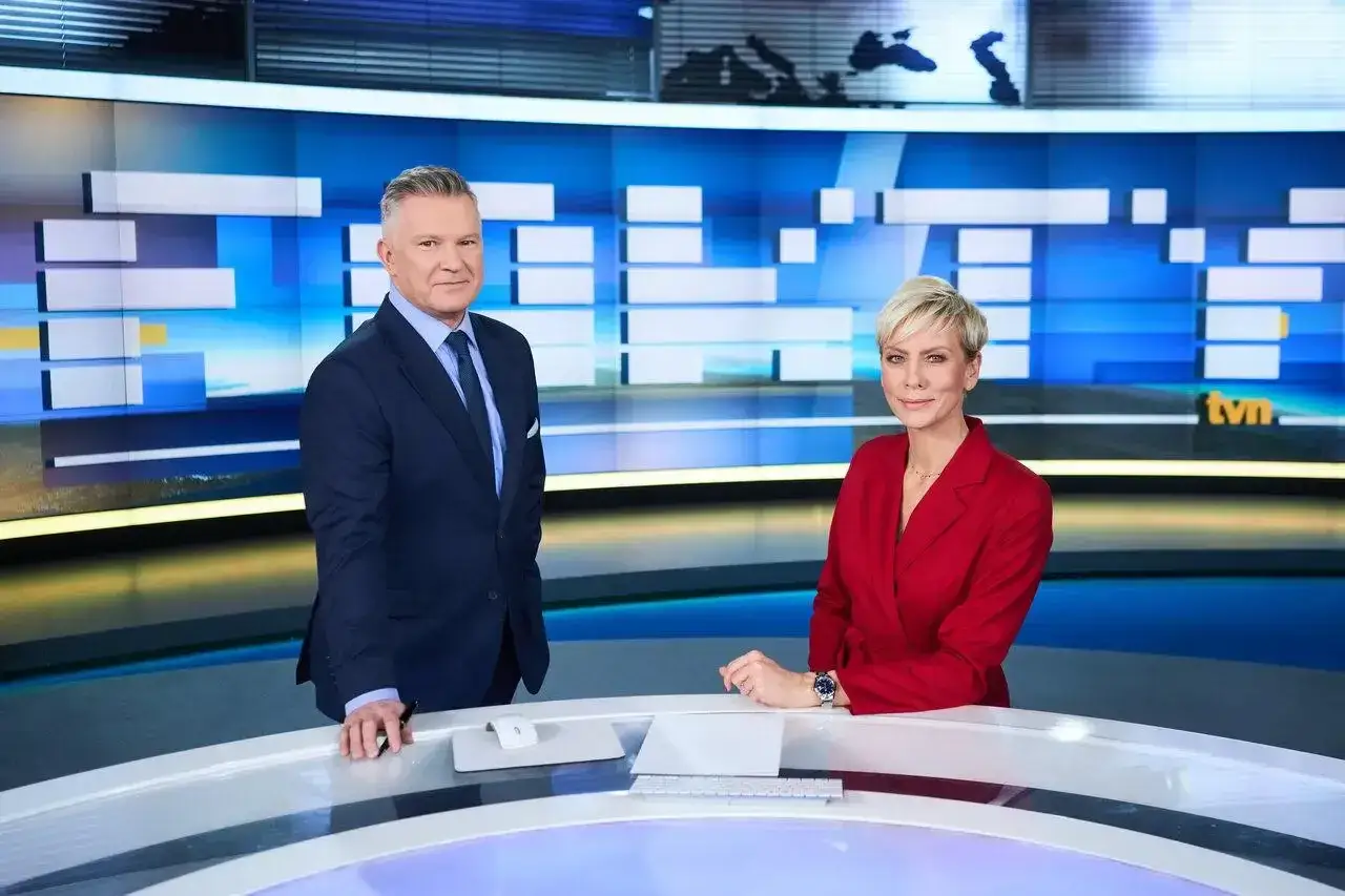 Prezenterzy Fakty TVN, mężczyzna w granatowym garniturze i kobieta w czerwonej marynarce, stoją przy stole w studiu telewizyjnym.