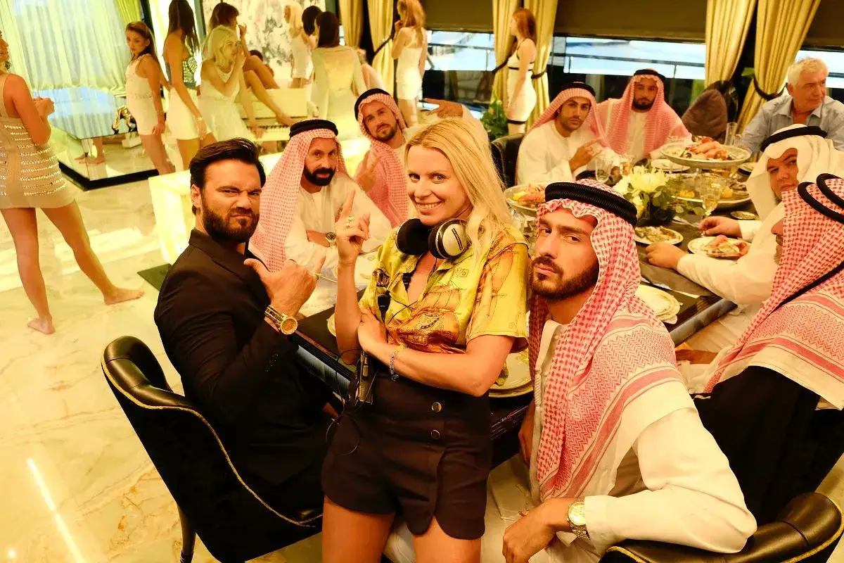 Grupa ludzi, w tym mężczyźni w tradycyjnych arabskich strojach i kobiety, siedzi przy stole. To scena z filmu 