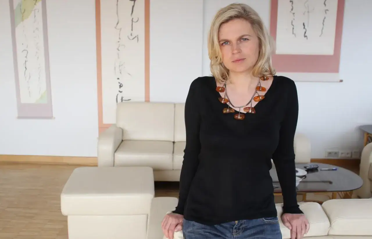 Barbara Nowacka, w wieku, gdy dzieci dorastają, w czarnej bluzce i z bursztynowym naszyjnikiem, siedzi na kanapie.