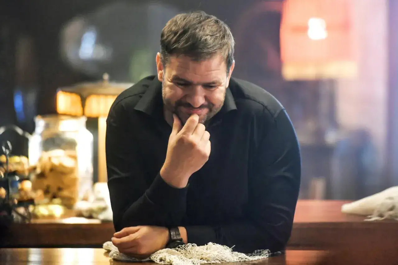 Tomasz Karolak wiek: aktor zamyślony, oparty o blat, w czarnej koszuli.