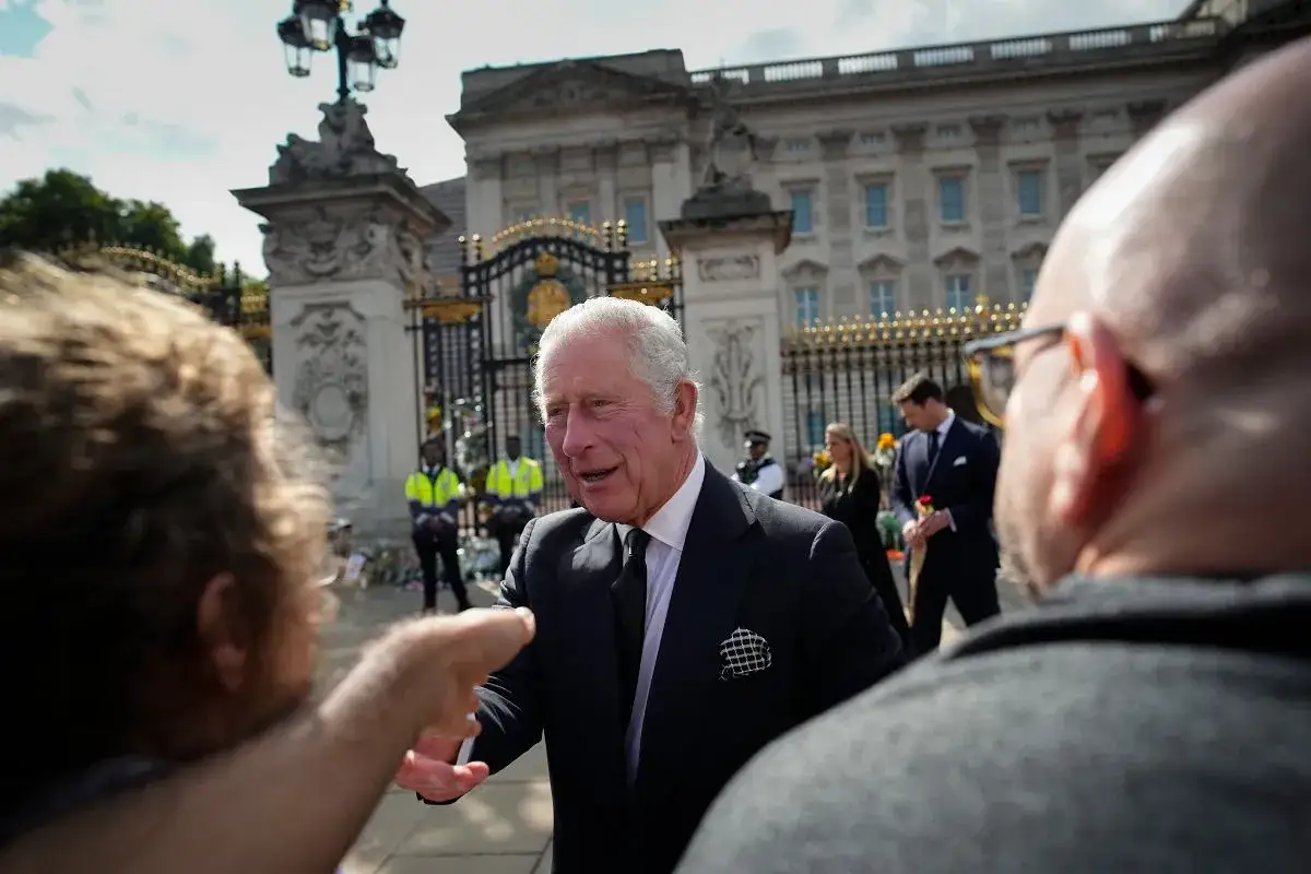 Król Karol w wieku, w garniturze, rozmawia z ludźmi przed Pałacem Buckingham.