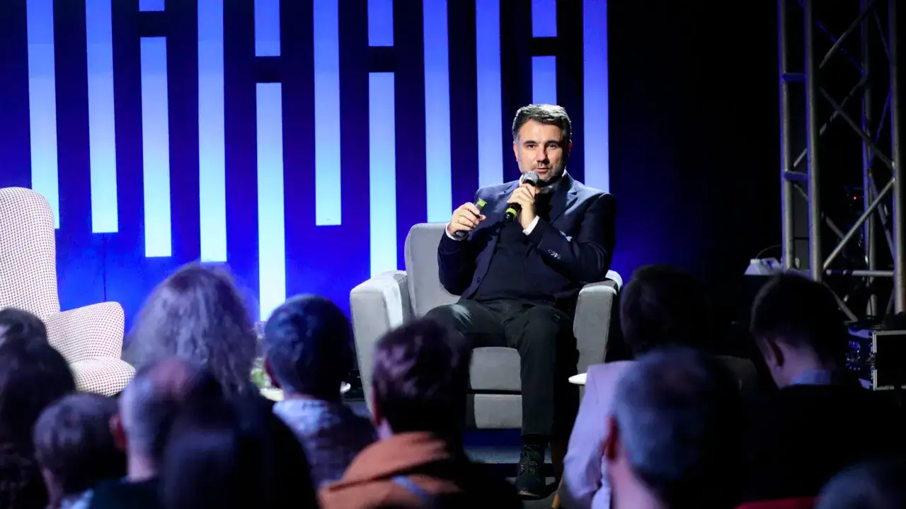 Marcin Majewski z TVN przemawia do publiczności, siedząc na wygodnym fotelu na tle niebieskiej scenografii.