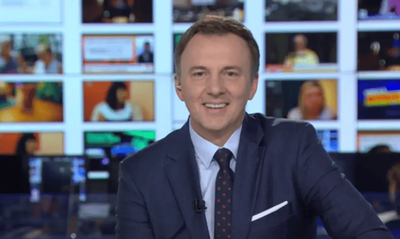 Krzysztof Górlicki w garniturze, uśmiechnięty, na tle ekranów.