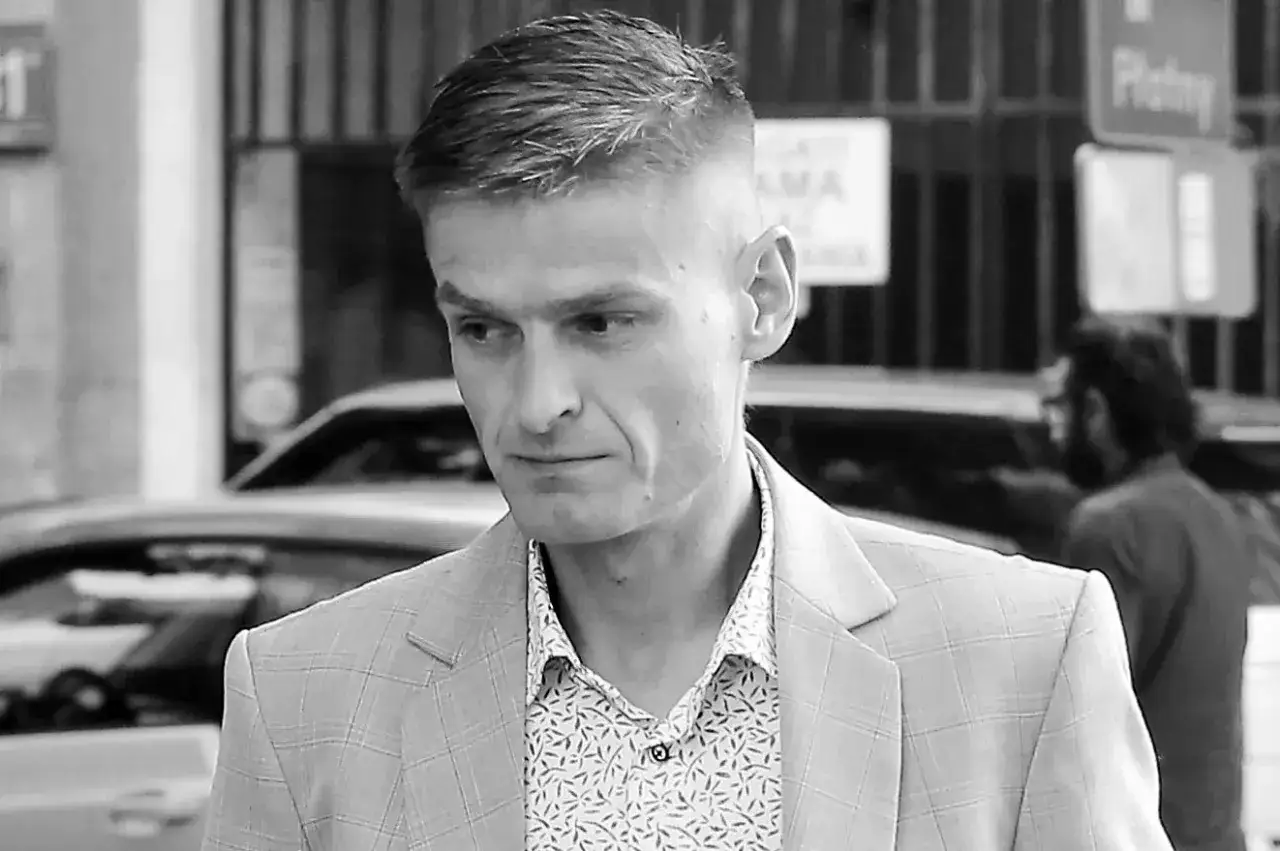 Tomasz Komenda, w wieku około 30 lat, w jasnej marynarce w kratę i wzorzystej koszuli, spogląda w bok.