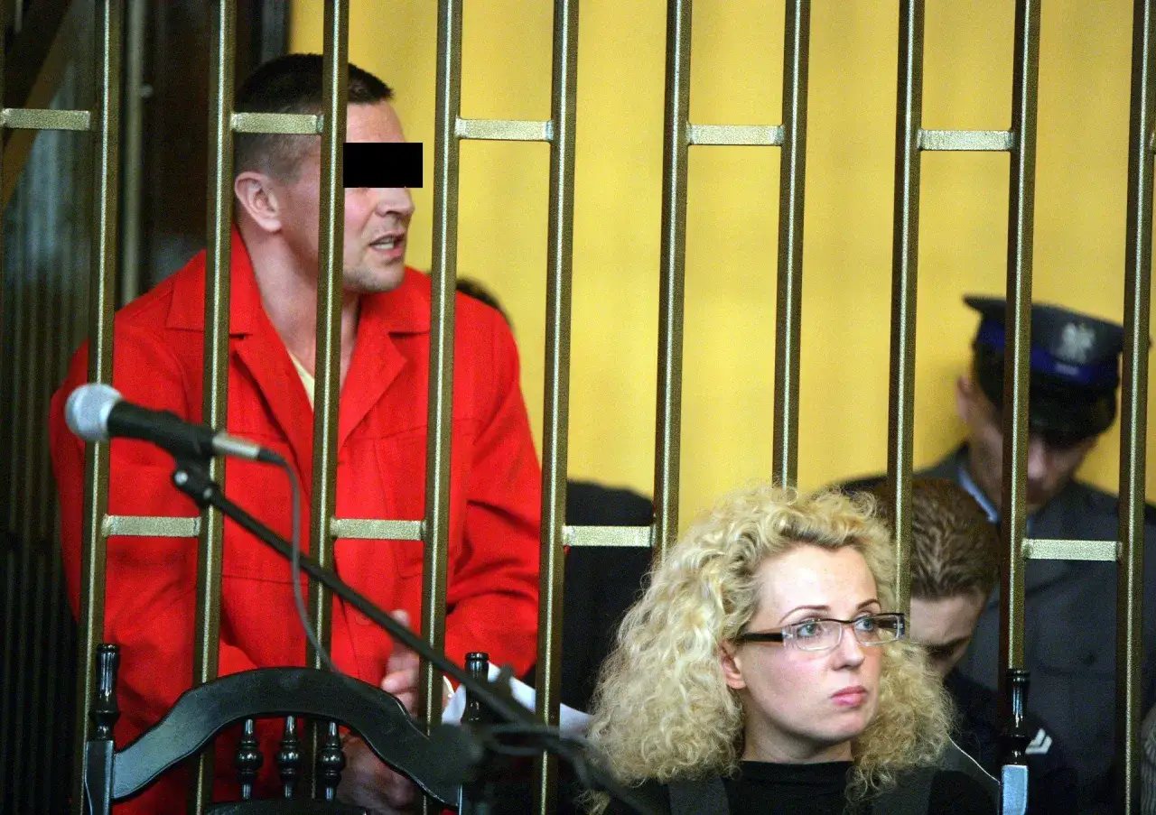 Mężczyzna w czerwonym ubraniu, przypominający lelek gangster, stoi za kratami. Obok siedzi kobieta w okularach.