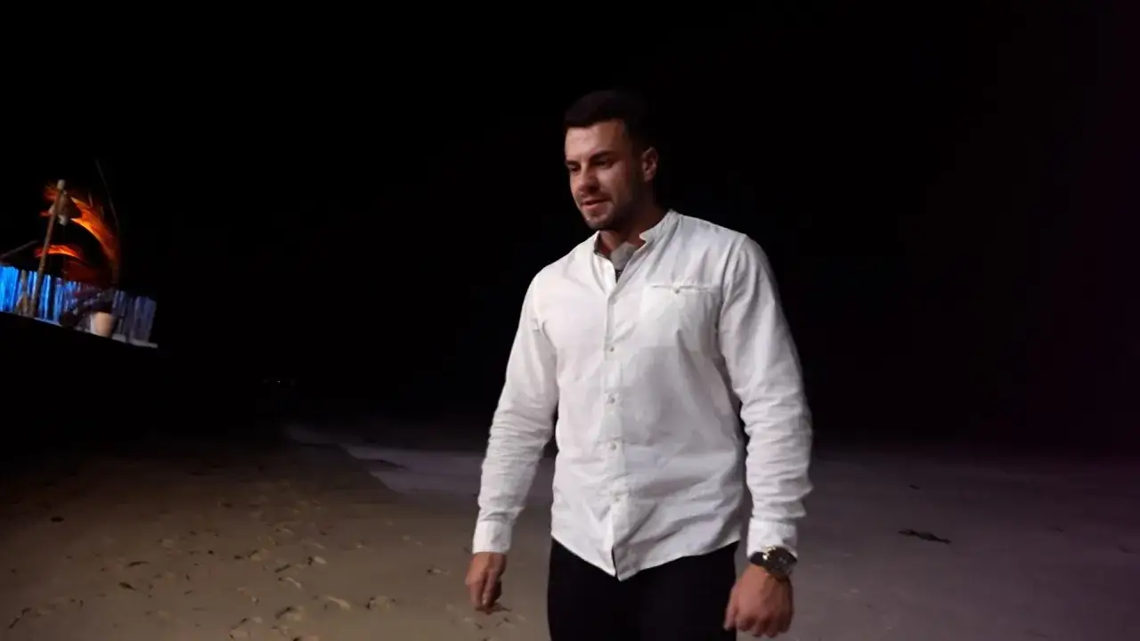 Mężczyzna w białej koszuli na plaży nocą. W tle widać ozdobną konstrukcję z ogniem, przypominającą statek. To może być scena z Kris Hotel Paradise.