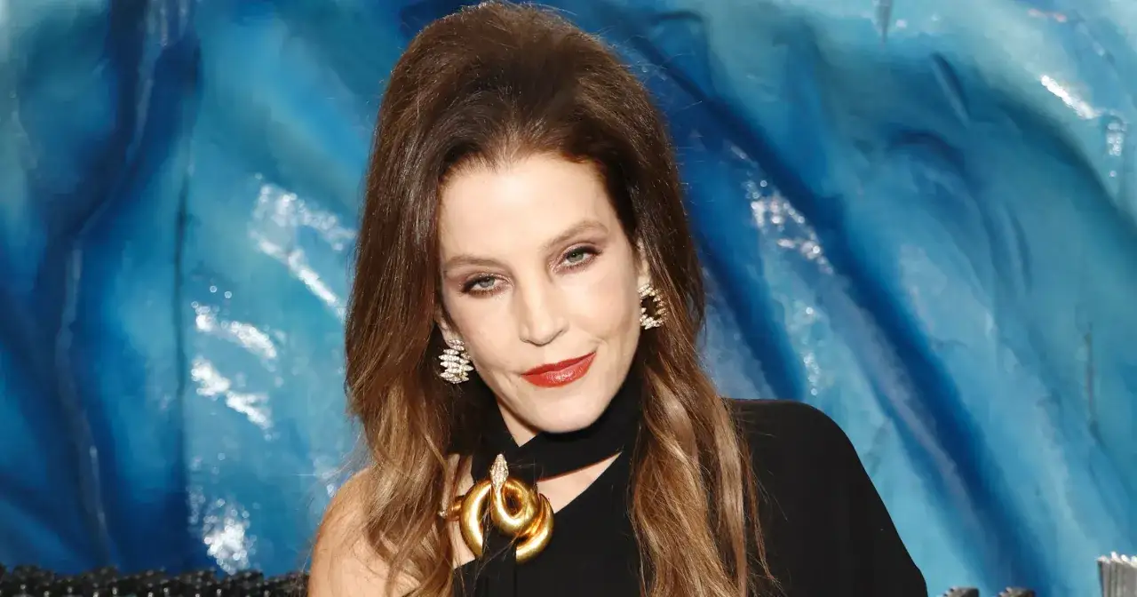 Lisa Marie Presley w szpitalu, z błyszczącymi kolczykami i złotym naszyjnikiem w kształcie węża, na tle niebieskiej, falującej tkaniny.