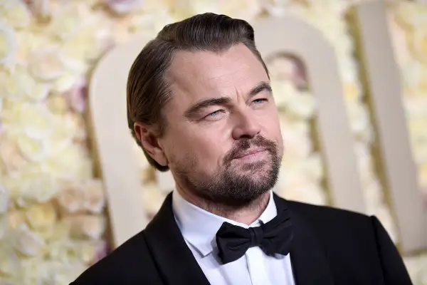 Leonardo DiCaprio wiek: aktor w czarnym smokingu z muszką, z siwiejącymi włosami i zarostem, na tle kwiatowej dekoracji.